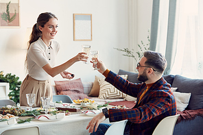 Portrait of beautiful young woman clinking glasses with husband across elegant dining table set for Christmas celebration, copy space