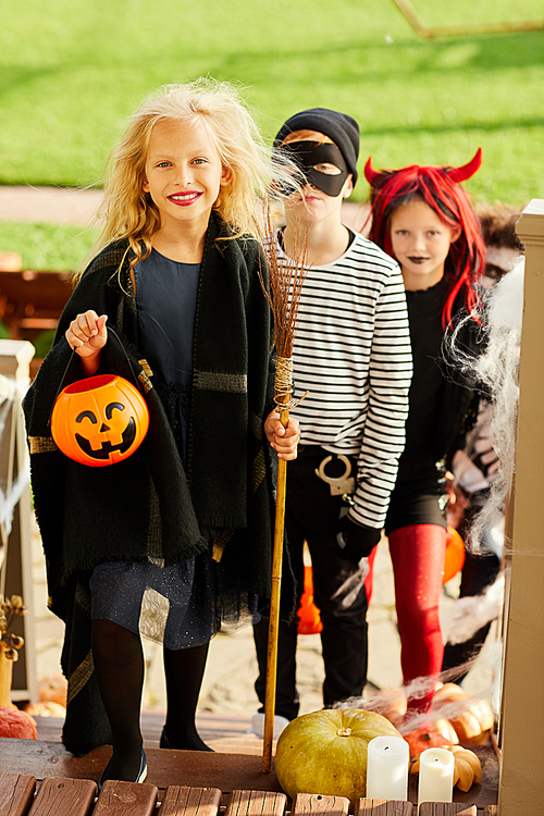 Full length portrait of children trick or treating on Halloween standing on stairs in row, focus on happy girl holding pumpkin basket