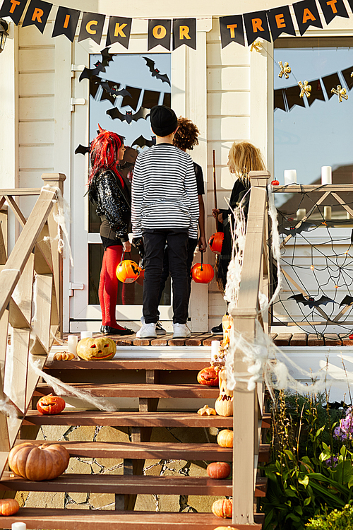 Back view of children trick or treating on Halloween, kids standing on porch knocking on doors of decorated house