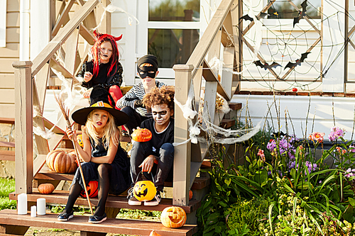 Multi-ethnic group of children wearing Halloween costumes  while sitting on stairs of decorated house in trick or treat season, copy space