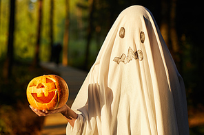 Waist up portrait of funny little kid dressed as ghost holding pumpkin while posing outdoors on Halloween, copy space