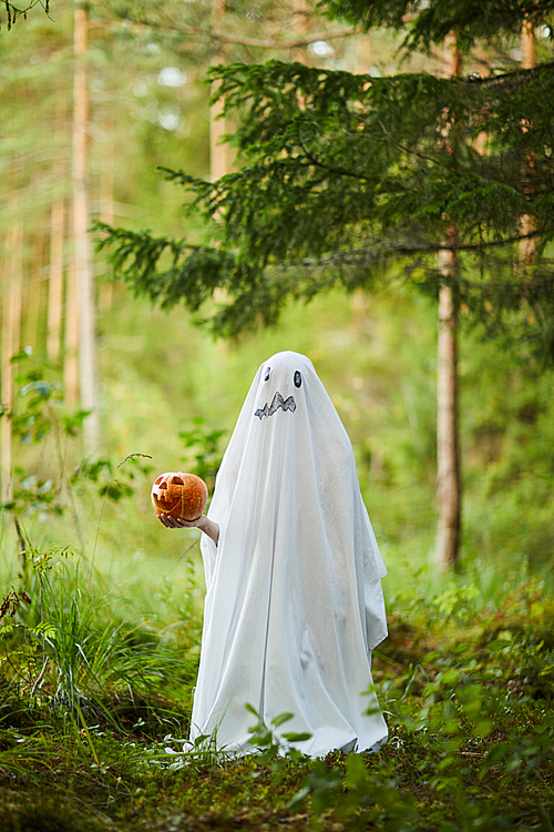 Full length portrait of spooky child dressed as ghost holding pumpkin while posing in forest on Halloween, copy space