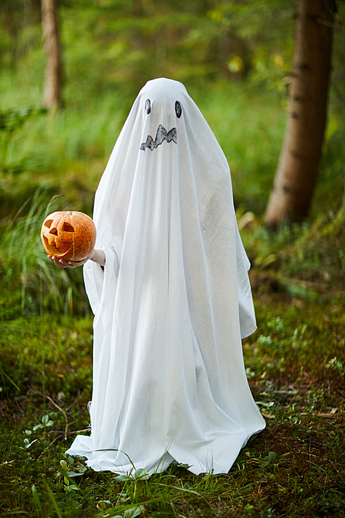 Full length portrait of spooky child dressed as ghost holding pumpkin while posing in forest on Halloween