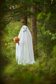 Full length portrait of spooky child dressed as ghost holding pumpkin standing in forest on Halloween