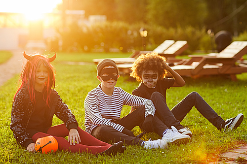 Full length portrait multi-ethnic group of kids wearing Halloween costumes sitting on green lawn outdoors and  lit by sunlight, copy space