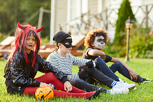 Full length portrait multi-ethnic group of kids wearing Halloween costumes sitting on green lawn outdoors, copy space