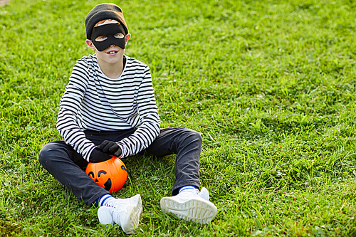 Full length portrait of cute boy wearing Halloween costume  while sitting on green grass with trick or treat basket, copy space