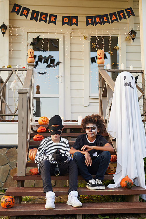 Portrait of two teenage boys wearing Halloween costumes sitting on porch of decorated house and , copy space