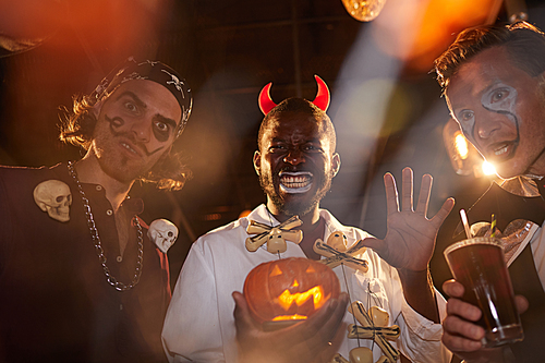 Group of adult men wearing Halloween costumes posing during party in club, focus on African-American man holding pumpkin in center