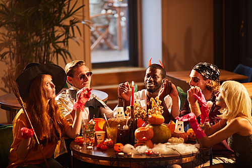 Group of friends wearing Halloween costumes performing magic ritual sitting round table at party, copy space
