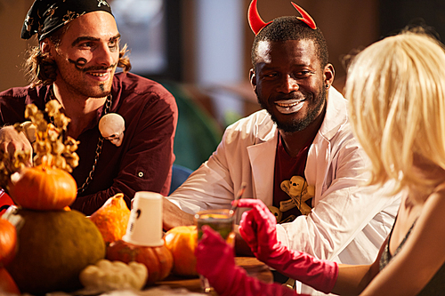 Multi-ethnic group of friends wearing costumes enjoying Halloween party sitting round table together, copy space