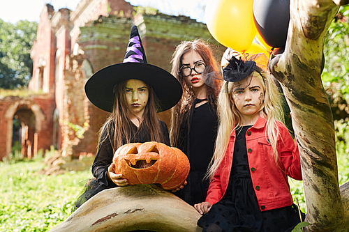 Staged portrait of three girls wearing Halloween costumes posing outdoors with pumpkin and balloons, copy space