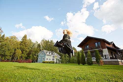 Wide angle portrait of happy little witch flying on broom above green lawn in Halloween, copy space