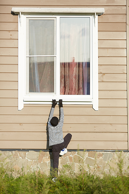 Full length portrait of unrecognizable little robber climbing to window of house on sunny day, copy space
