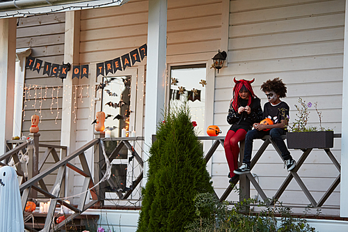 Wide angle view at boy and girl eating Halloween candy sitting on fence after trick or treating together, copy space