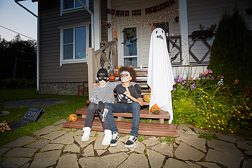 Full length portrait of two boys wearing Halloween costumes posing outdoors sitting on porch together in dark, shot with flash, copy space