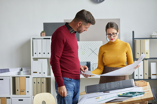 Portrait of two mature architects man and woman working on blueprints together while standing by drawing desk in office, copy space