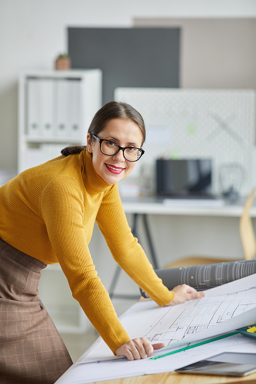 Vertical portrait of adult female architect smiling at camera while leaning on drawing desk in office and working on blueprints