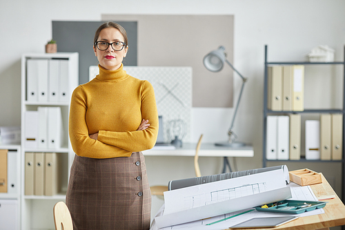 Waist up portrait of confident female architect  while standing with arms crossed by drawing desk in office, copy space