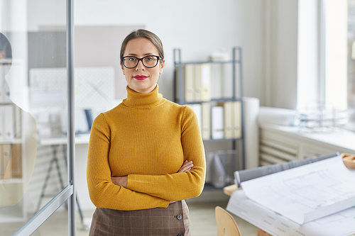 Waist up portrait of confident female architect  and smiling while standing with arms crossed by drawing desk in office, copy space