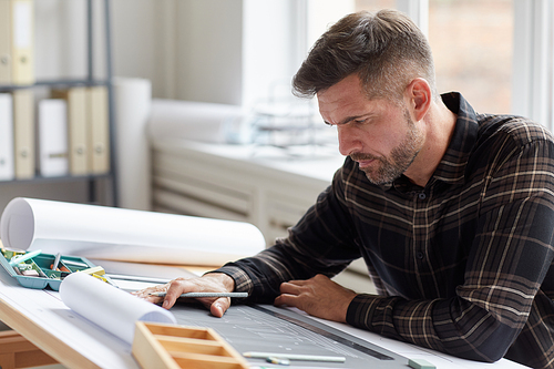 Portrait of mature bearded architect working on blueprints and plans while sitting at drawing desk in office, copy space
