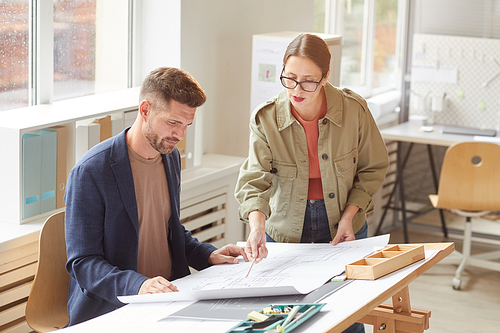 Waist up portrait of two architects pointing at blueprints and discussing work while standing by drawing desk in office lit by sunlight , copy space