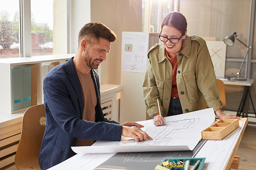 Waist up portrait of two smiling architects pointing at blueprints and discussing work while standing by drawing desk in office lit by sunlight , copy space