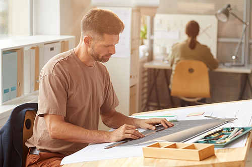 Side view portrait of mature bearded architect looking at blueprints while sitting at drawing desk in sunlight, copy space