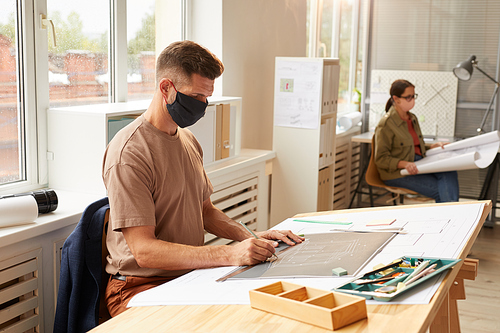 Side view portrait of mature bearded architect wearing mask while sitting at drawing desk in sunlight, copy space