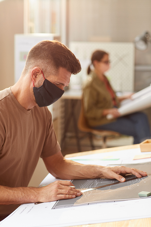 Side view portrait of mature bearded architect wearing mask while sitting at drawing desk in sunlight