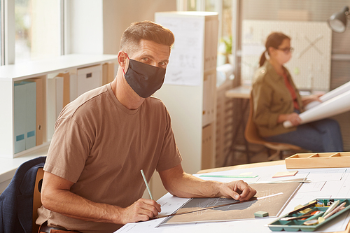 Portrait of mature bearded architect wearing mask while sitting at drawing desk in sunlight, copy space