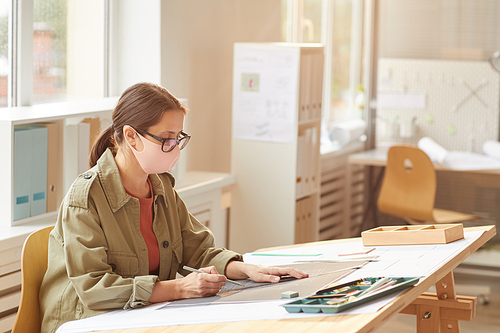 Side view portrait of female architect wearing mask while sitting at drawing desk in sunlight, copy space