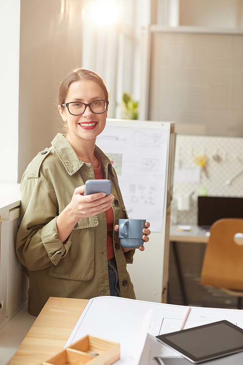 Vertical warm toned portrait of young woman enjoying coffee and holding smartphone during coffee break in office