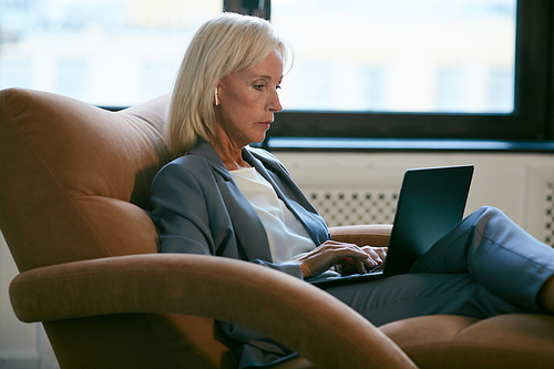 Side view portrait of modern mature businesswoman working with laptop while siting in leather chair in office, copy space