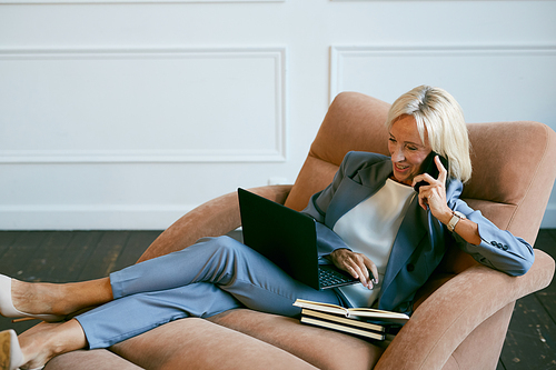 Portrait of smiling mature businesswoman speaking by smartphone while working in lounge chair in office, copy space