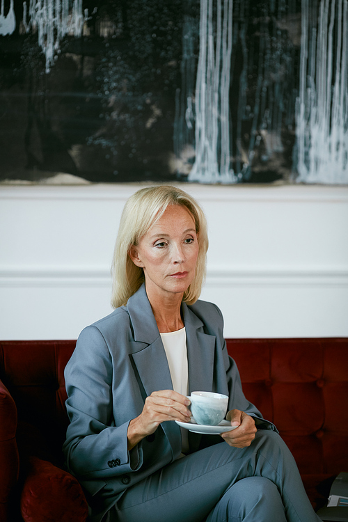 Vertical portrait of modern mature businesswoman holding coffee cup and looking away pensively while sitting on velvet sofa in cafe