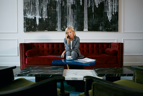 Wide angle portrait of modern mature businesswoman speaking by phone while sitting on velvet sofa in cafe, copy space