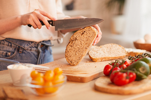 Warm-toned close up of unrecognizable woman cutting fresh wholewheat bread while making breakfast in cozy kitchen, copy space