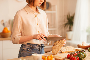 Mid-section portrait of modern young woman cutting fresh wholewheat bread while making breakfast in cozy kitchen, copy space