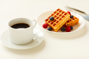 Warm-toned close up of sweet dessert waffles with berry topping next to cup of black coffee on white background, copy space