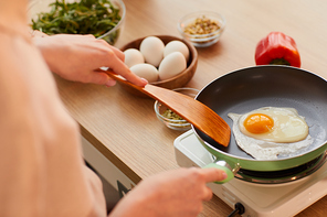 Warm-toned close up of unrecognizable young woman cooking eggs on frying pan while preparing healthy breakfast in morning, copy space
