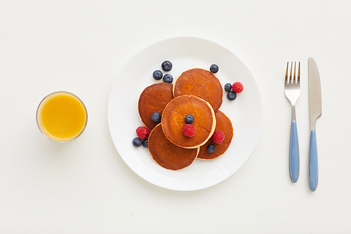 Top view at minimal composition of delicious golden pancakes with fresh berries next to orange juice and knife and fork on white background, breakfast concept, copy space