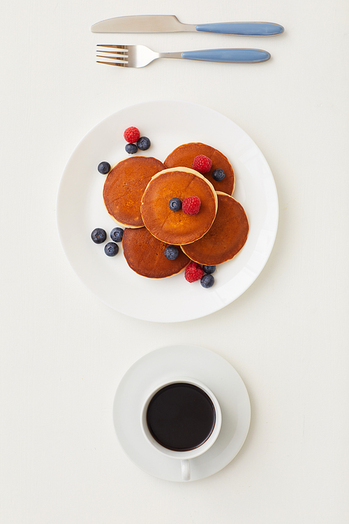 Top view at minimal composition of delicious golden pancakes decorated with fresh berries next to coffee cup on white background, breakfast concept, copy space