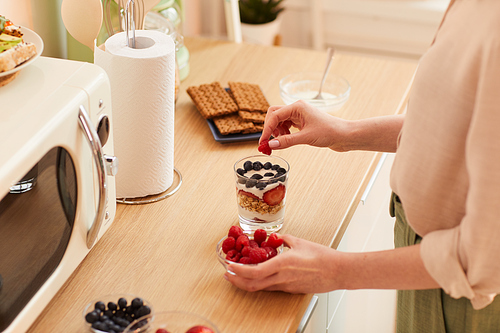 Warm-toned close up of unrecognizable young woman making yogurt parfait with fresh berries and granola while preparing healthy breakfast in morning, copy space