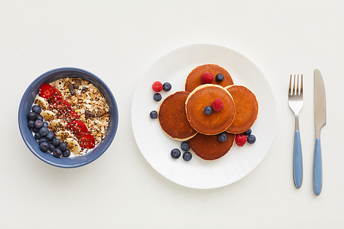 Top view at minimal composition of delicious golden pancakes decorated with fresh berries next to granola plate on white background, healthy breakfast concept, copy space