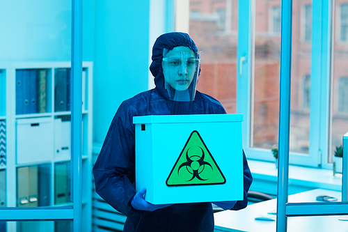 Waist up portrait of male scientist wearing biohazard gear holding box with danger sign while working on research in bio laboratory, copy space