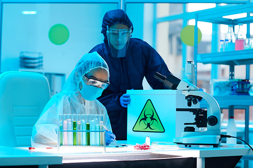Portrait of two scientists wearing biohazard gear holding box with danger sign while working on research in bio laboratory, copy space
