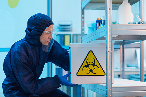 Side view portrait of male scientist wearing biohazard gear holding box with danger sign while working on research in bio laboratory, copy space