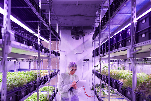 Wide angle portrait of female agricultural engineer writing on clipboard while standing between shelves in plant  greenhouse lit by blue light, copy space