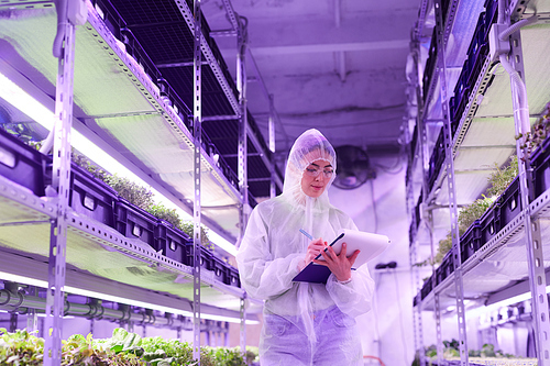 Portrait of female agricultural engineer writing on clipboard while standing between shelves in plant  greenhouse lit by blue light, copy space